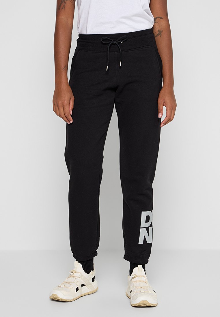 Persona che indossa pantaloni jogger neri con testo bianco su una gamba, sneakers bianche, calze bianche e una t-shirt bianca infilata nei pantaloni.