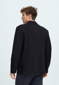 Junger Mann mit lockigem Haar, der ein lockeres schwarzes Blazer trägt, nach links blickt und von hinten sowie von der Seite sichtbar ist, vor einem schlichten Hintergrund.
