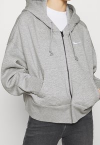 Grå zip-up hoodie gjord av mjukt tyg, med en huva med dragsko, framfickor och en liten vit logotyp på bröstet.