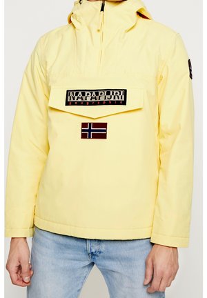 Veste jaune à capuche avec poche avant, logo Napapijri et écusson du drapeau norvégien, portée avec un jean bleu clair.