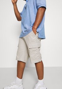 Camisa polo azul claro, pantalones cortos tipo cargo beige con bolsillos laterales, y zapatillas blancas con detalles en negro. Tejido de algodón y corte relajado.