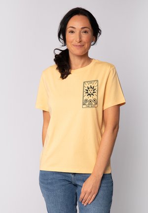 Geel katoenen t-shirt met korte mouwen, met een zwart zon grafisch ontwerp op de voorkant, gecombineerd met blauwe spijkerbroeken.