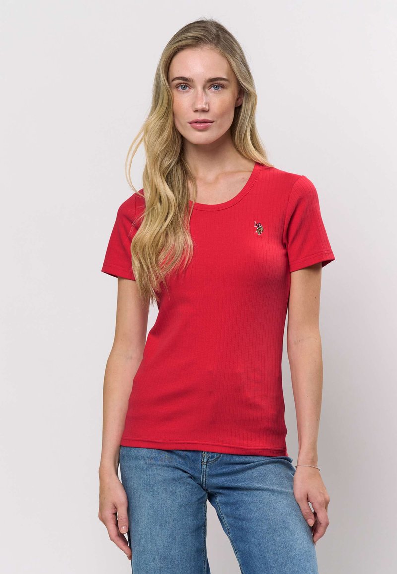 Rotes, geripptes T-Shirt mit kurzen Ärmeln, rundem Halsausschnitt und kleinem gesticktem Akzent auf der Brust. Kombiniert mit blauen Jeans.