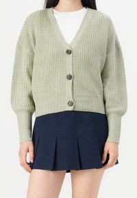 Jasnozielony dzianinowy kardigan z dekoltem w serek, pięcioma guzikami oraz prążkowanym dołem i mankietami, w połączeniu z granatową plisowaną spódnicą.