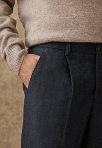 Pantalons en laine à chevrons bleu marine avec poches latérales, présentant un devant plat et une taille standard. Accompagné d'un pull beige en maille côtelée.