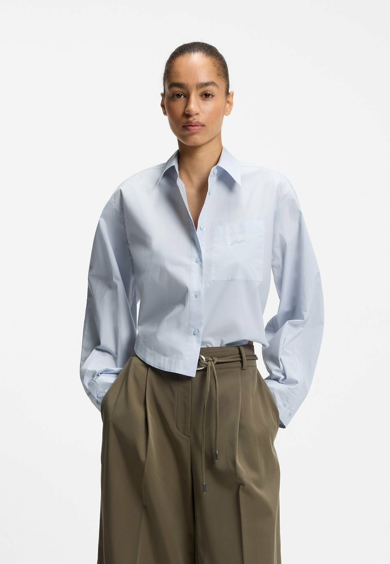 Femme portant une chemise courte bleu clair boutonnée et un pantalon large taille haute vert olive, mains dans les poches.