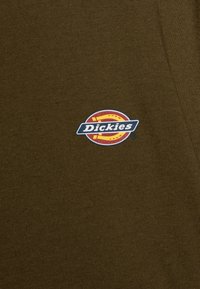 Olivově zelená látka má vyšitou nášivku Dickies, kruhový design s červenými a žlutými akcenty na námořnickém pozadí. Jednoduché texturované pozadí.