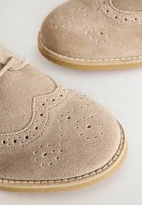 Chaussures en daim beige avec des détails de brogue, présentant des motifs perforés et une semelle en caoutchouc marron clair contrastante. Design de lacets taillés.