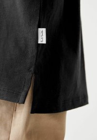 Crna kratka majica s kratkim rukavima s bočnim prorezima, izrađena od mekanog materijala, s bijelom etiketom na kojoj piše "Paul Smith" u kurzivu.