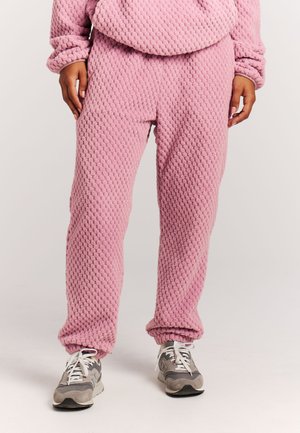 Persoon die een roze joggingbroek met textuur draagt, met een bijpassende top en grijze sneakers, staand tegen een effen lichte achtergrond.