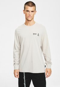 Jasnobeżowa koszulka z długim rękawem i okrągłym dekoltem, z małym czarnym logo na piersi oraz ściągaczami przy mankietach. Wykonana z miękkiej bawełny.