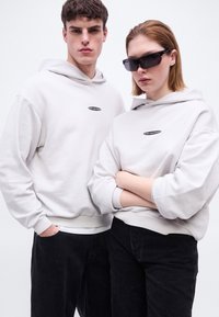 Νεαρός άνδρας και γυναίκα φορώντας ταιριαστές λευκές κουκούλες και μαύρο παντελόνι, η γυναίκα με κοντά μαλλιά φοράει μαύρα γυαλιά ηλίου, ποζάρουν μπροστά σε λευκό φόντο.