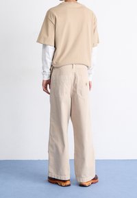 Camiseta de algodón beige superpuesta sobre una camisa de manga larga blanca, combinada con pantalones beige holgados con bolsillos traseros, usados con zapatillas multicolores.