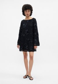 Melna kleita ar apaļu kakla izgriezumu, garām zvana piedurknēm un ziedu izšuvumiem. Puscaurspīdīga auduma, līdz ceļiem, ar nedaudz izplestu siluetu.