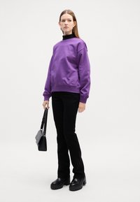 Felpa viola con polsini a coste, pantaloni neri aderenti, chunky neri con plateau e una piccola borsa nera. Sfondo uniforme.
