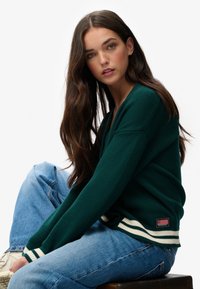 Groene gebreide trui met wit en marineblauwe gestreepte manchetten, met een decoratieve patch aan de zoom. Gecombineerd met lichtblauwe spijkerbroek.