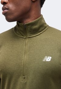 Άνδρας που φοράει πράσινο ελιά quarter-zip πουλόβερ με λευκό λογότυπο New Balance στο στήθος, κοιτάζει αριστερά.