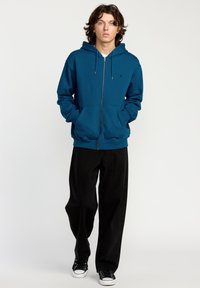 Sudadera con cremallera en color verde azulado, hecha de un material suave, con un bolsillo frontal y capucha con cordones, combinada con pantalones amplios negros y zapatillas negras.