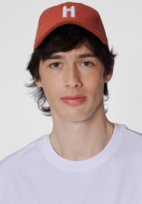 Röd baseballkeps med vit "H"-logotyp, bärs �över kort mörkt hår; enkel vit t-shirt med rund halsringning; neutral bakgrund.