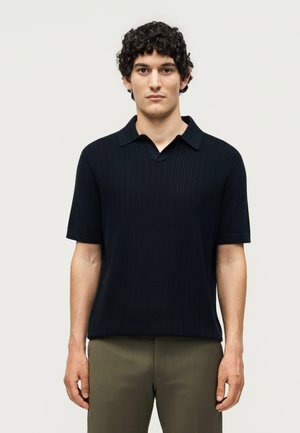 Homme aux cheveux bouclés portant un polo en maille bleu marine à manches courtes et un pantalon vert olive, debout devant un fond clair uni.