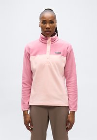 Γυναίκα που φοράει ροζ και ανοιχτό ροζ fleece μπλούζα Columbia με κουμπιά τύπου snap, στέκεται μπροστά σε λευκό φόντο.