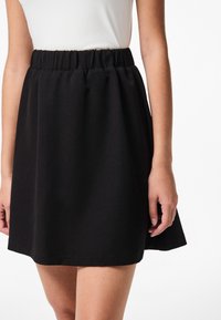 Žena u crnoj suknji s elastičnim pojasom uparena s bijelom topićem bez rukava, kratkom do sredine bedra, stoji ispred jednobojne bijele pozadine.