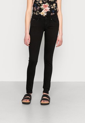 Slim-fit sorte jeans båret med sort blomster ærmeløs top og sorte sandaler med dobbelt rem på lys grå gulv og hvid væg baggrund.