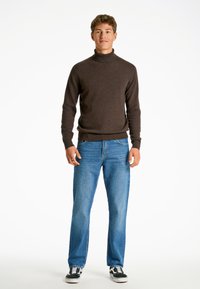 Brun polotröja i ett mjukt material, kombinerad med ljusblå raka jeans och svarta sneakers, stående mot en vit bakgrund.