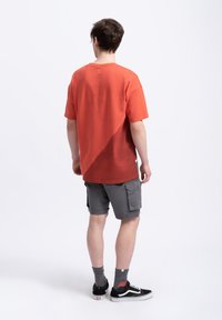 T-shirt en coton orange à manches courtes, présentant un motif de dégradé diagonal, associé à un short cargo gris et des baskets noires.