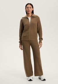 Maglione marrone a zip con colletto alto, abbinato a pantaloni larghi coordinati. Il materiale presenta un motivo strutturato. Scarpe bianche completano l'outfit.