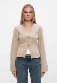 Beige cardigan laget av myk strikket stoff, med V-hals, frontlukking med knapper og brede ermer med knappeaksenter. Båret over en hvit topp.