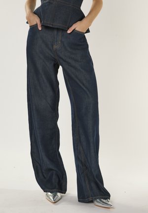 Pantaloni in denim blu scuro a gamba larga con cuciture a contrasto, dotati di tasche frontali e una texture morbida. Abbinati a scarpe metallizzate con punta affilata.