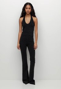 Hosszú sötét hajú nő, fekete ujjatlan nyakpántos topban és hozzá illő bő szabású nadrágban áll, egy sima fehér háttér előtt.