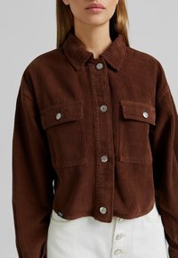 Hnědá kordurová bunda s límcem, dvěma náprsními kapsami, stříbrnými knoflíky a krátkým střihem. Hladká textura s vertikálními pruhy.
