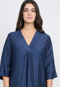 Marineblaue Satinbluse mit V-Ausschnitt, kurzen Ärmeln und Faltendetail in der Mitte. Glatte Textur mit einem leichten Glanz.