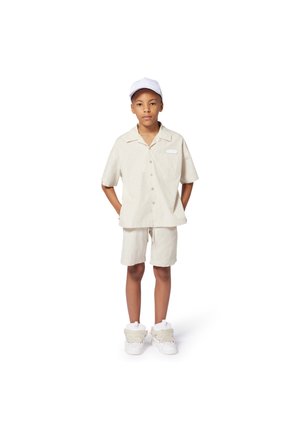 Chemise beige clair à manches courtes avec un col et une poche avant, assortie à un short. Une casquette blanche et des baskets blanches chunky complètent le look.