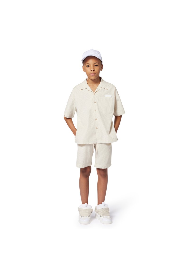 Licht beige overhemd met korte mouwen, een kraag en een borstzak, gecombineerd met bijpassende shorts. Een witte pet en chunky witte sneakers maken het geheel af.