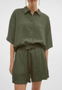 Camicia verde oliva, a maniche corte, con colletto e chiusura a bottone. Abbinata a pantaloni corti con vita elastica dello stesso colore, dotati di cordoncino.