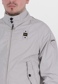Giacca zip-up grigia chiara con collo alto, dettagli in bottoni e patch con logo sulla spalla. Realizzata in materiale liscio e leggero.