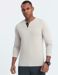 Man i beige långärmad henley-tröja med svart knappslå och mörk armbandsur, som står mot en enkel ljus bakgrund.