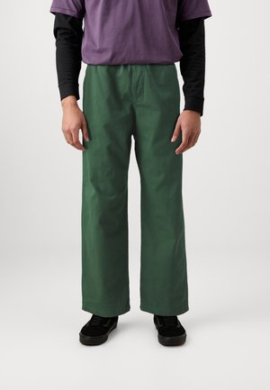 Green wide-leg pants made of cotton blend fabric, featuring a smooth texture and a relaxed fit. Paired with a purple and black layered top.