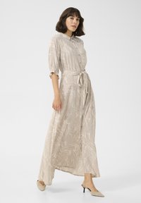 Robe longue de couleur crème avec un motif marbré, manches courtes bouffantes, devant à boutons, taille cintrée et ceinture nouée. Portée avec des talons beiges.