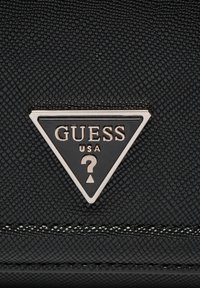 Materiale nero testurizzato con un prominente logo triangolare nero e argento "GUESS" e un emblema di punto interrogativo sulla parte anteriore.