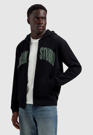 Man draagt een zwarte zip-up hoodie met de tekst "STUDIO" op de borst, wit overhemd eronder en blauwe spijkerbroek, kijkt naar rechts.