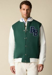 Man in een groen-wit varsity jack met "BB" patch, witte geribbelde manchetten en drukknoopsluiting, gecombineerd met crèmekleurige broek.