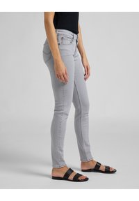 Jeans ajustados en gris claro con una cintura de altura media, bolsillos delanteros y detalles de costura negra. Combinados con sandalias planas negras.