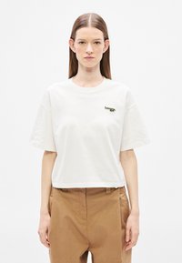 Camiseta corta blanca de algodón con mangas cortas, con un bordado de logo verde en el lado izquierdo. Textura suave.