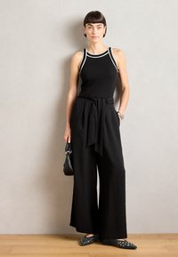 Top sin mangas negro con detalles blancos, combinado con pantalones negros de pierna ancha y cintura alta. Accesorios incluyen un bolso pequeño negro y zapatos planos de punta.