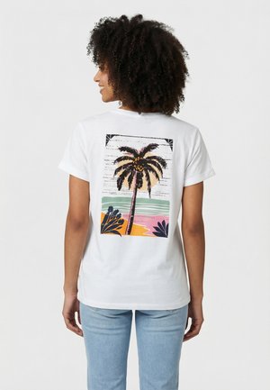 Donna con capelli ricci che indossa una maglietta bianca con un design colorato di palme e paesaggio astratto sul retro, abbinata a jeans azzurri.