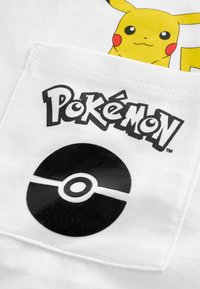Hvid bomulds t-shirt med Pikachu grafik ovenfor "Pokémon" logoet og et sort Poké Ball design med blank overflade på lommeområdet.